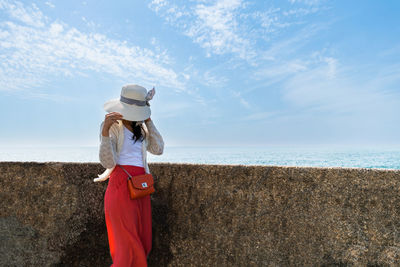 Woman wearing a cardigan sweater is hiding behind hat with a blue sea and sky background