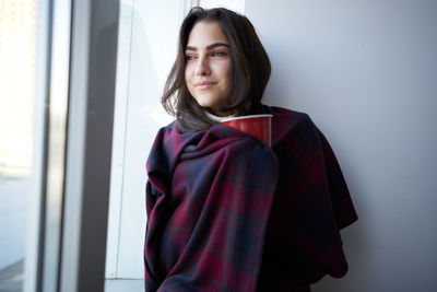 Young woman wrapped in blanket standing against wall at home