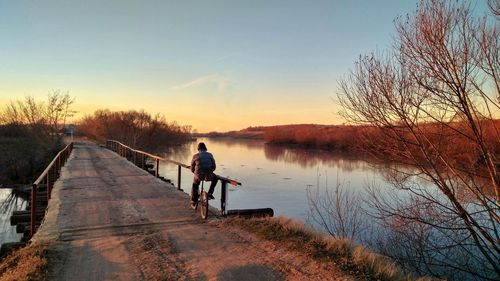 Rear view of man riding bicycle on footbridge over lake during sunset