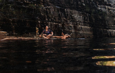 Man swimming in lake while woman sitting by rock formation in forest