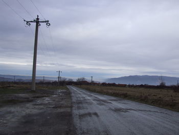 Road by electricity pylons on field against sky