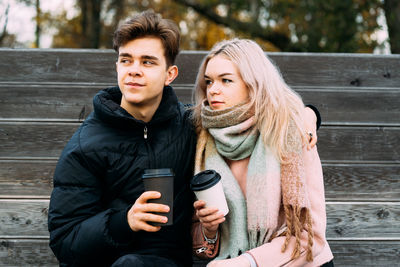 Young couple holding disposable coffee cups while sitting on bench at park during autumn