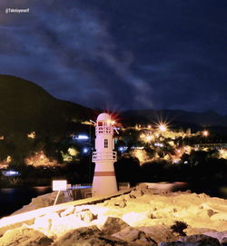 Illuminated building by lighthouse against sky at night