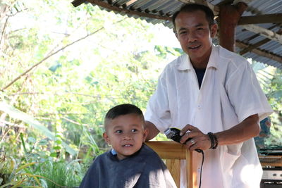 Portrait of mature barber trimming boy hair at shop against trees