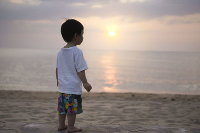 Rear view of a little boy on the beach standing looking at the vast sea against sky during sunset. 