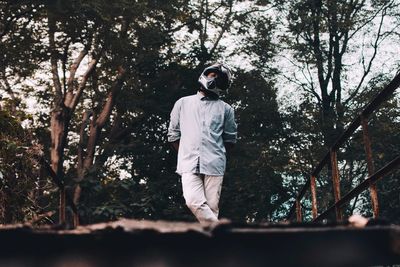 Low angle view of young man standing on footbridge against trees in forest