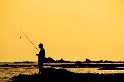 Silhouette man fishing at beach against orange sky