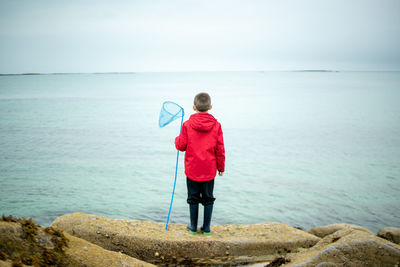 Rear view of boy with fishing net looking at sea against sky