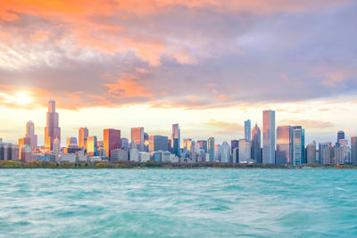 Sea by modern buildings against sky during sunset