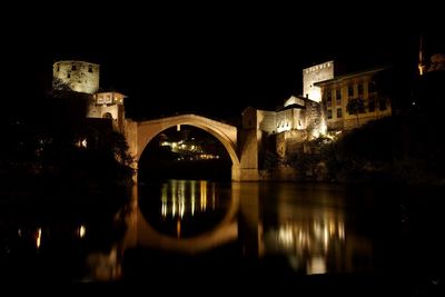 Illuminated arch bridge over river by buildings in city at night