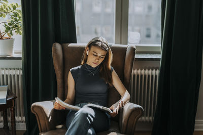 Teenage girl reading book while sitting on armchair against window at library