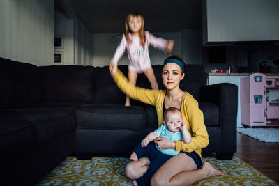 A young mother holding her baby while her toddler jumps on the couch