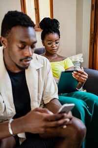 Content young african american lady reading information on tablet while resting on sofa with boyfriend using smartphone at home