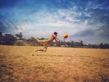 Full length of young man playing volleyball on sand at beach against cloudy sky