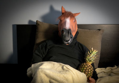 Man wearing horse mask while holding pineapple while lying on bed at home