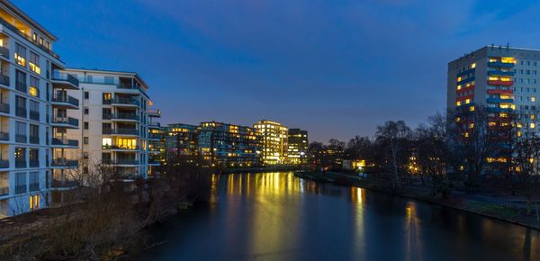 Illuminated buildings by river against sky at dusk