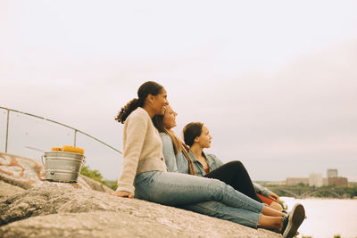 Happy female friends looking away while sitting on rock formation at lakeshore against sky
