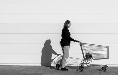 Side view of woman walking with shopping cart on footpath against wall