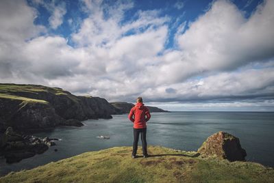 Rear view of man standing on cliff by sea against sky