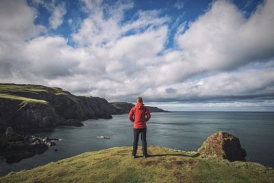 Rear view of man standing on cliff by sea against sky