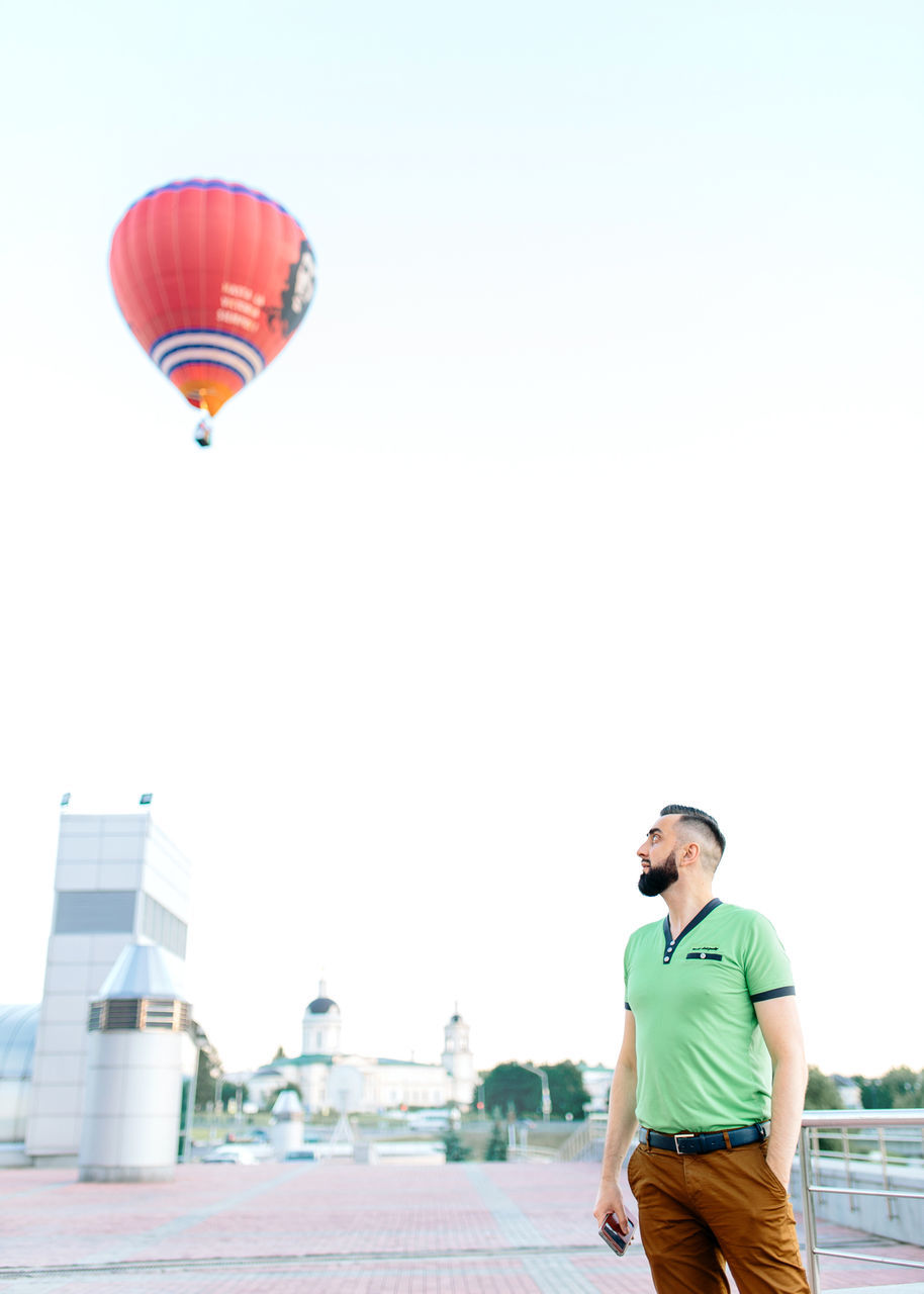 real people, leisure activity, one person, lifestyles, clear sky, outdoors, mid-air, built structure, young men, architecture, building exterior, sky, young adult, standing, day, hot air balloon, city