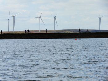 People riding bicycle on riverbank by windmills against sky