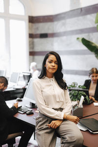 Portrait of confident female financial advisor sitting on desk in law office