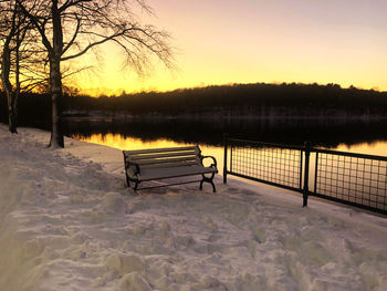 Bench by lake during winter against sky at sunset