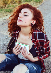 Young woman with eyes closed holding book while sitting on road