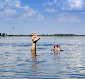 Cropped hands drowning in lake against sky