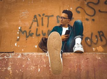 Full length of young man looking away while sitting against wall