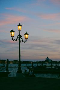 Illuminated street light by river against sky during sunset