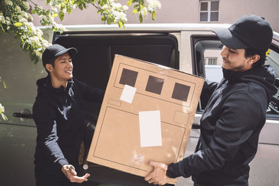 Smiling delivery men holding cardboard box near truck