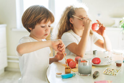 Two siblings brother and sister toddler boy tween girl painting easter eggs on kitchen at home 