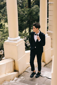 A beautiful young man, the groom in an elegant wedding suit, stands posing in the city's old park