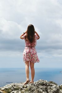 Full length rear view of woman looking at sea while standing on rock against cloudy sky