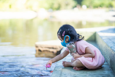 Cute girl wearing protective face mask feeding carp koi fishes at pond.