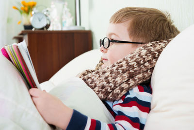 Side view of boy reading book while lying down on bed at home