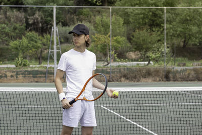 Portrait of young sporty boy with cap holding his tennis equipment and waiting to play on the court.