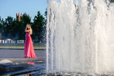 Young woman in pink evening gown standing by fountain in park