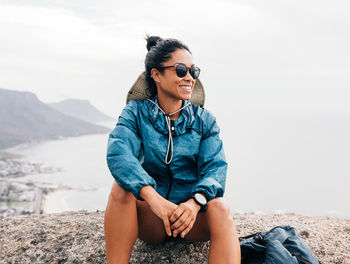 Young woman wearing sunglasses sitting on mountain under sky