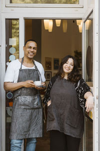 Portrait of happy multiracial male and female cafe owners standing at doorway