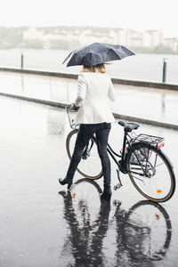 Full length rear view of businesswoman walking with bicycle on city street during rainy season