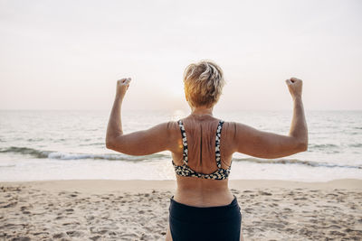 Rear view of senior woman flexing muscles while standing at beach