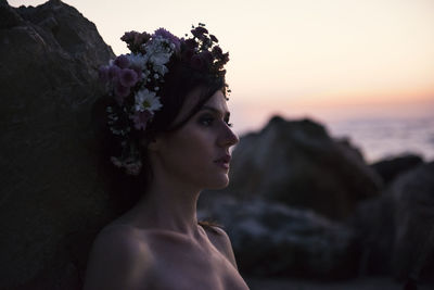 Woman wearing flowers while looking away against sky during sunset