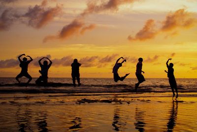 Silhouette friends jumping over beach against orange sky during sunset