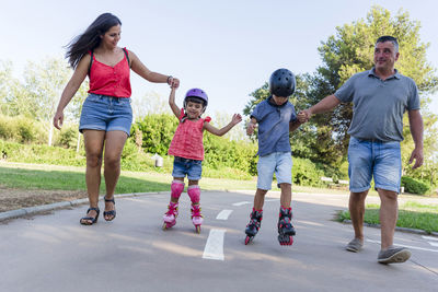 Kids holding her parents hands while roller skating outdoors in the park.