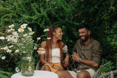 Happy friends talking while sitting on bench against plants in garden