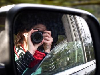 Reflection of woman on side-view mirror photographing through camera while sitting in car
