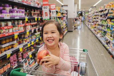Happy girl holding apple while sitting in shopping cart at store